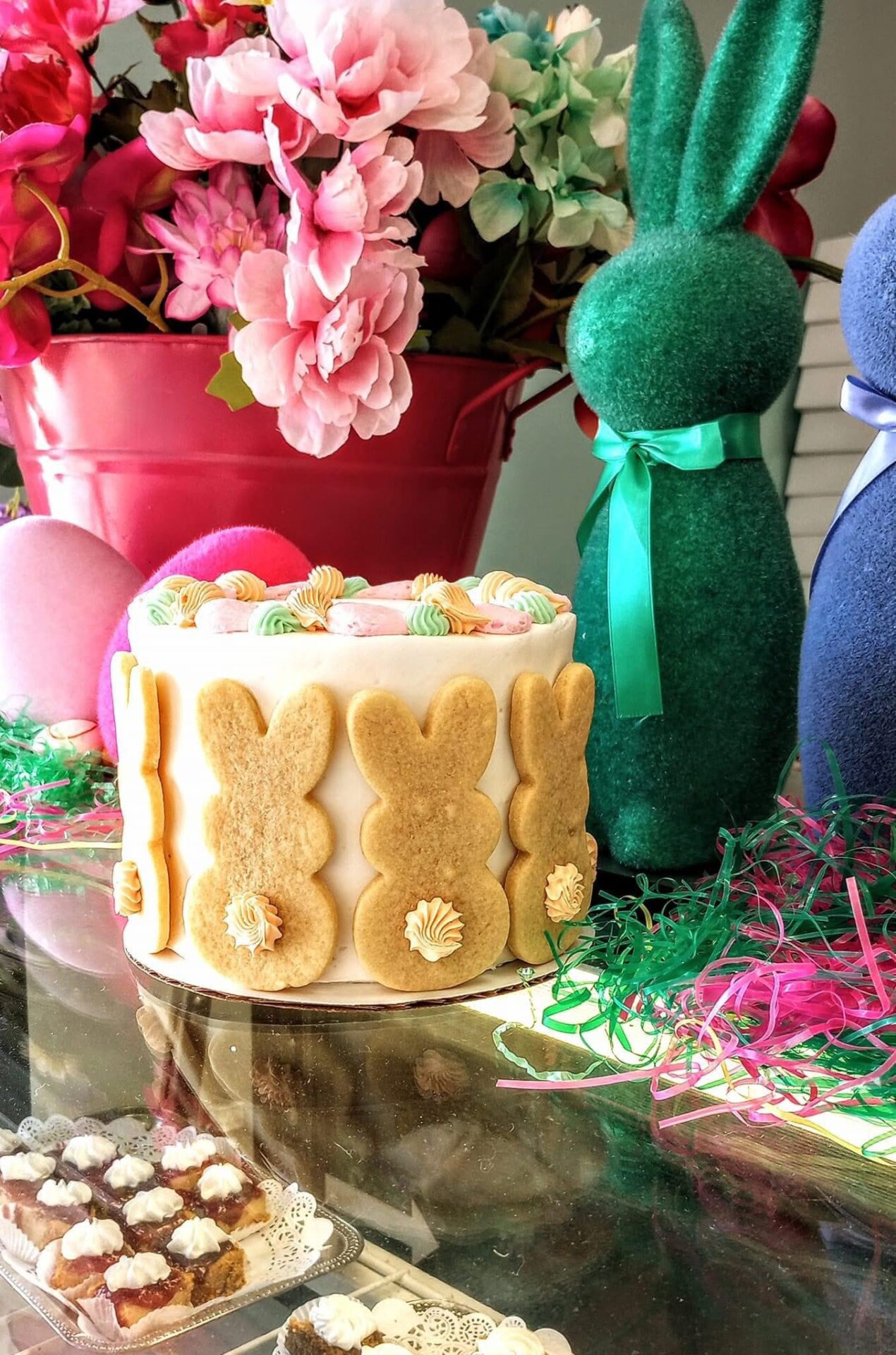 A festive Easter scene featuring a bunny-themed cake surrounded by colorful flowers, decorative eggs, and felt bunny figures